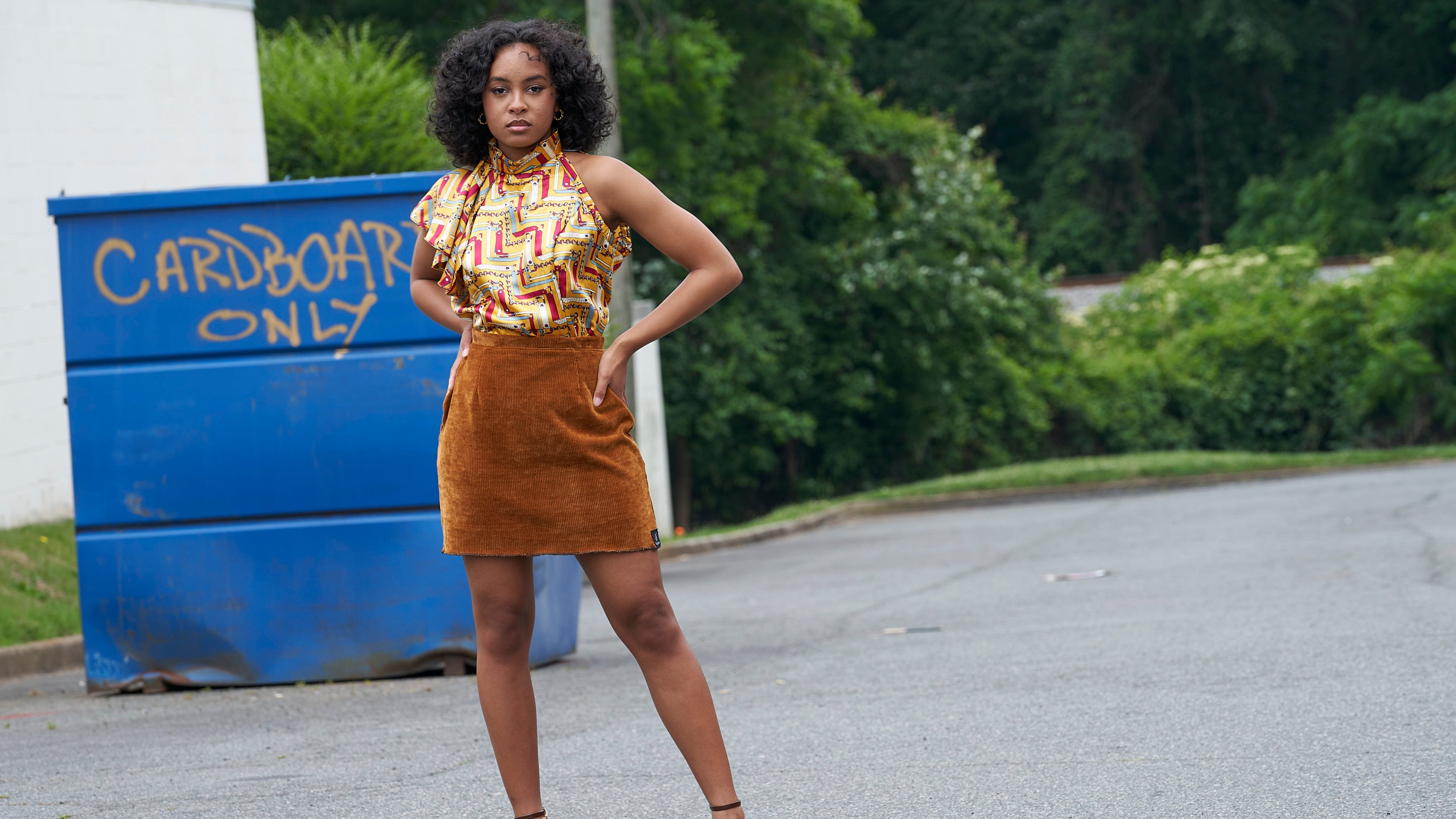 Woman in a patterned top and brown skirt standing on a road with a blue 'Cardboard Only' bin in the background.
